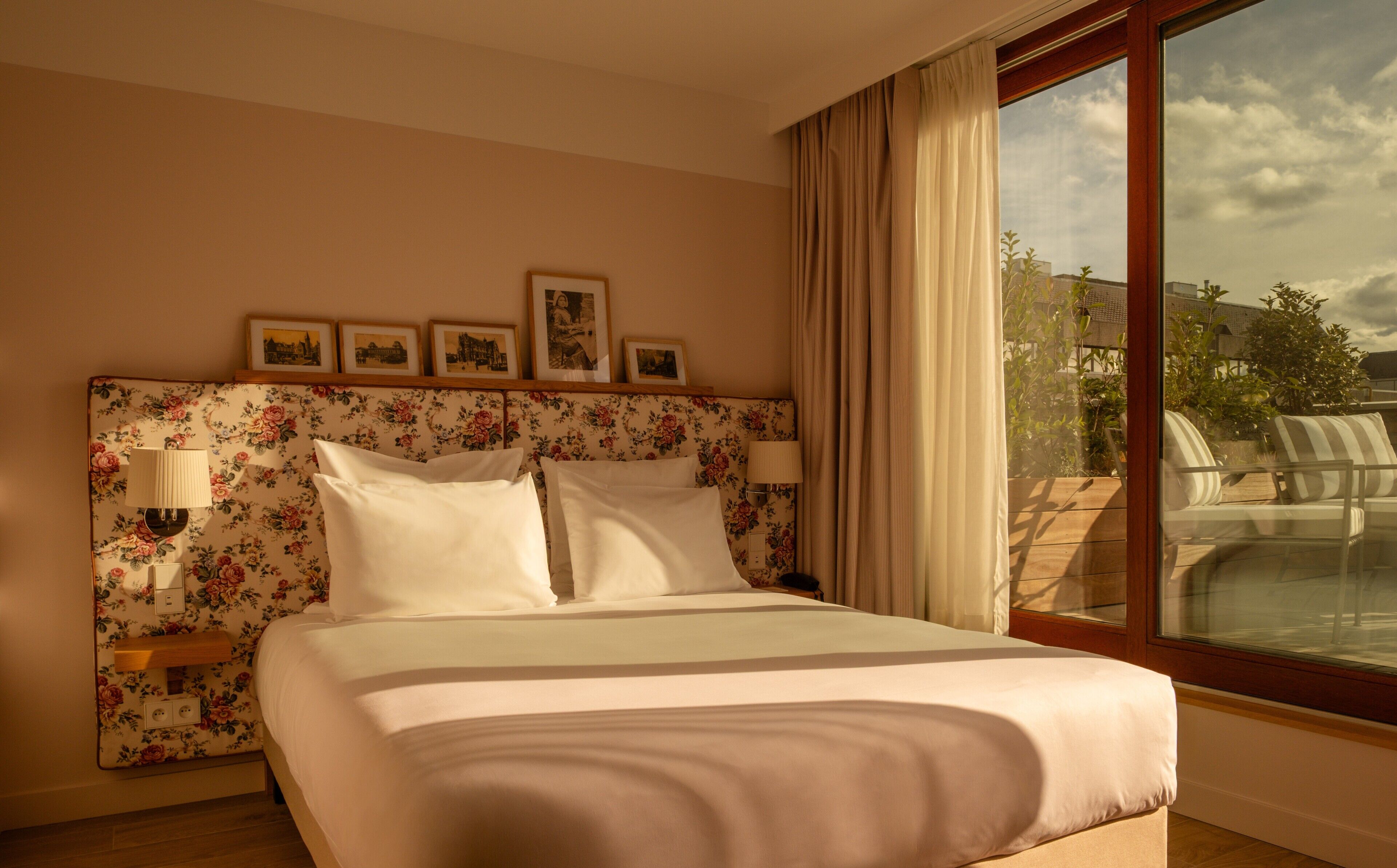 A hotel room with a floral headboard, a large bed, two bedside lamps, and a view of a pool through a large window.