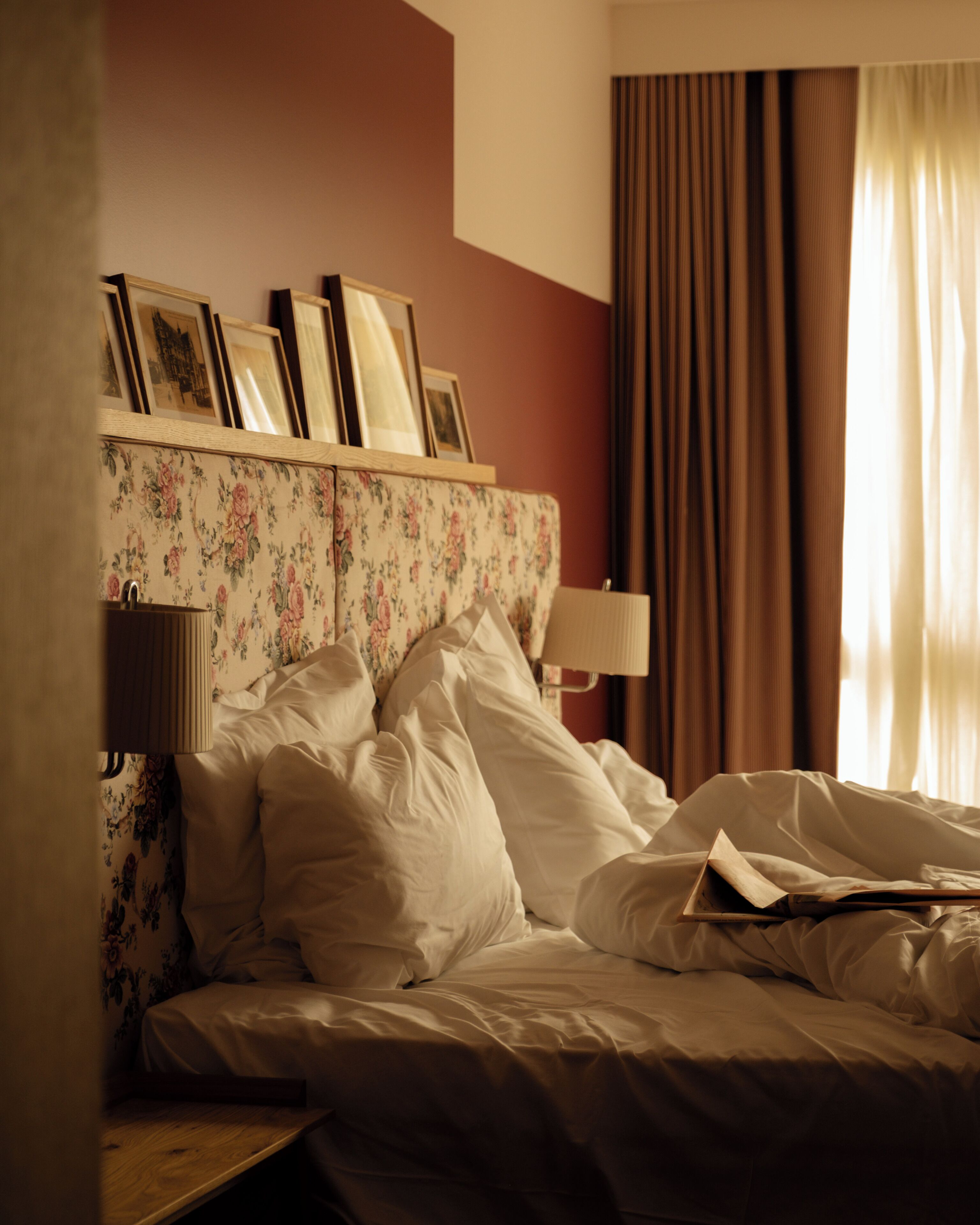 Habitación de hotel con cabecera de cama estampada con motivos florales, dos lámparas de noche, cama con ropa blanca y una ventana con cortinas.