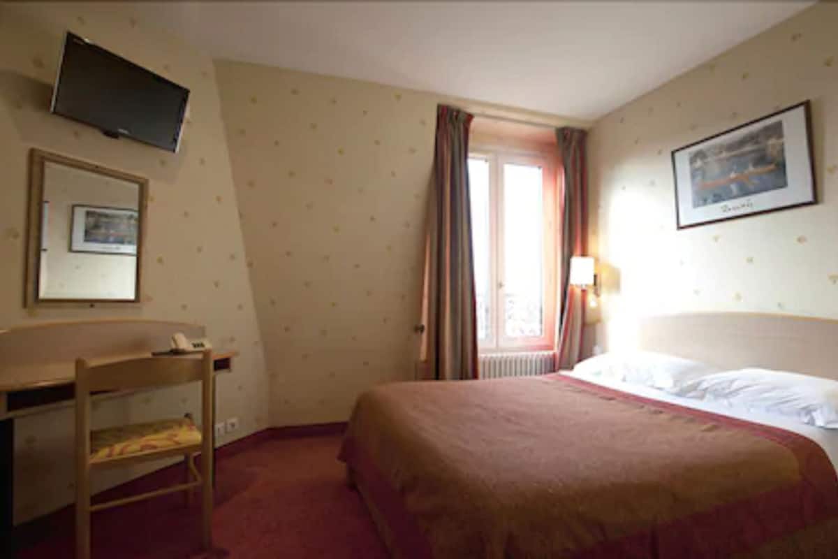 Ein Hotelzimmer mit Bett, Schreibtisch, Stuhl, Fernseher und einem Fenster mit Vorhängen.