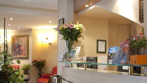 A hotel reception area with a glass counter, a red chair, potted plants, and a framed picture on the wall. - Hotel Transcontinental (Paris)