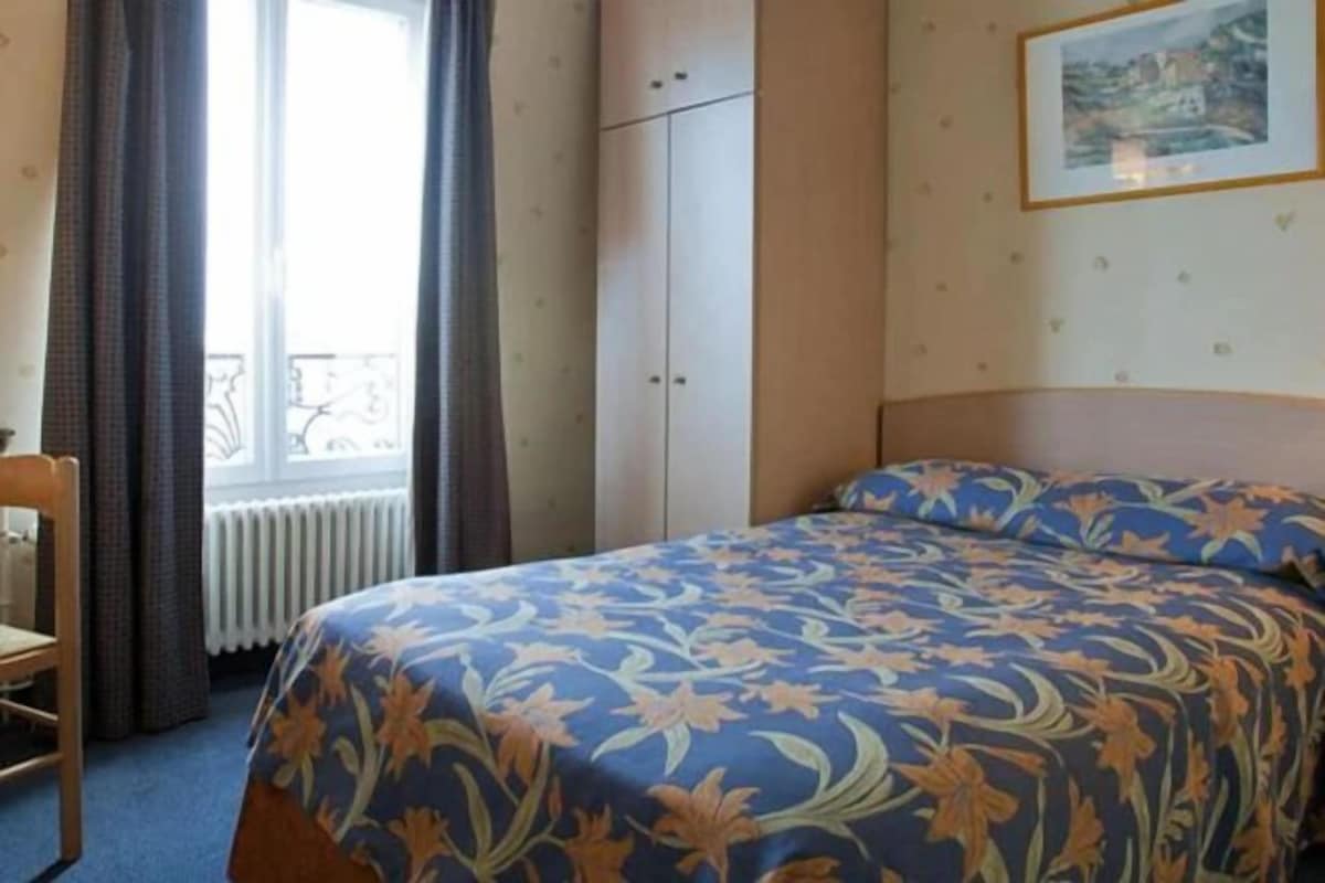 A hotel room with a bed, a chair, a window with curtains, a radiator, and a framed picture on the wall.