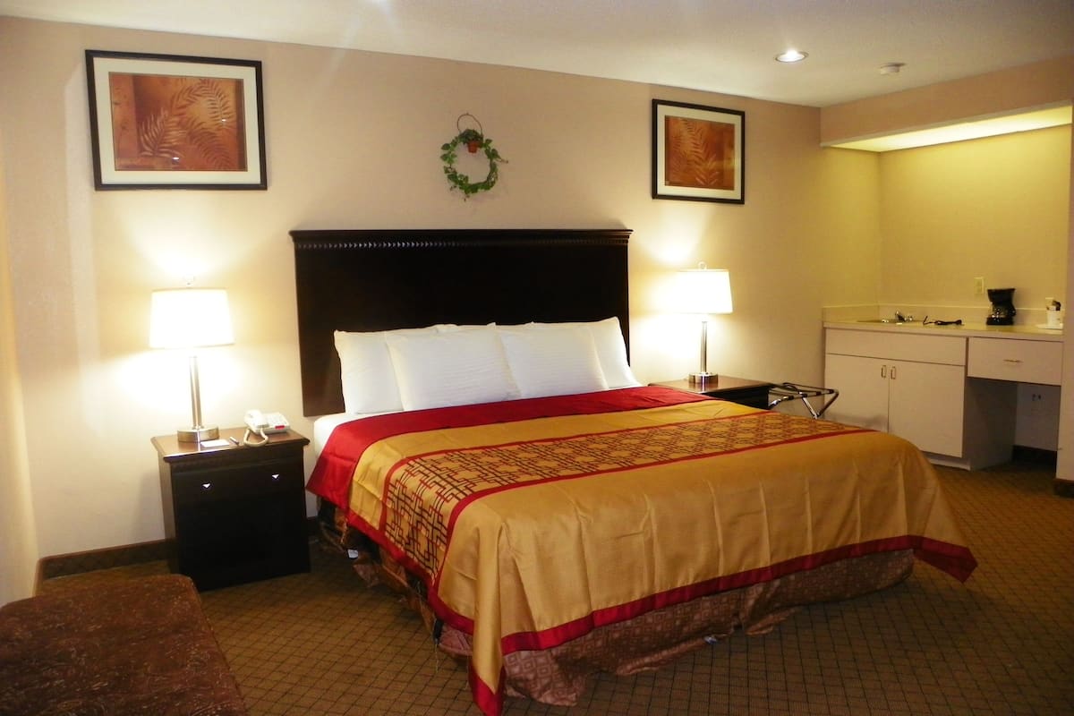 A hotel room with a large bed, two bedside lamps, a nightstand, a small table, a chair, a wall-mounted TV, a framed picture, and a kitchen area.