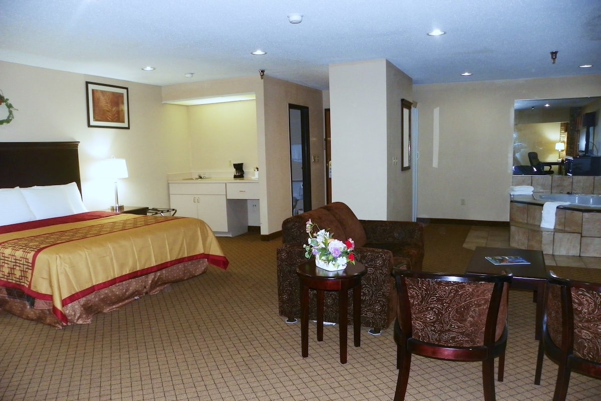 A hotel room with a bed, a desk, a chair, a small table with a vase, and a bathroom visible through an open door.