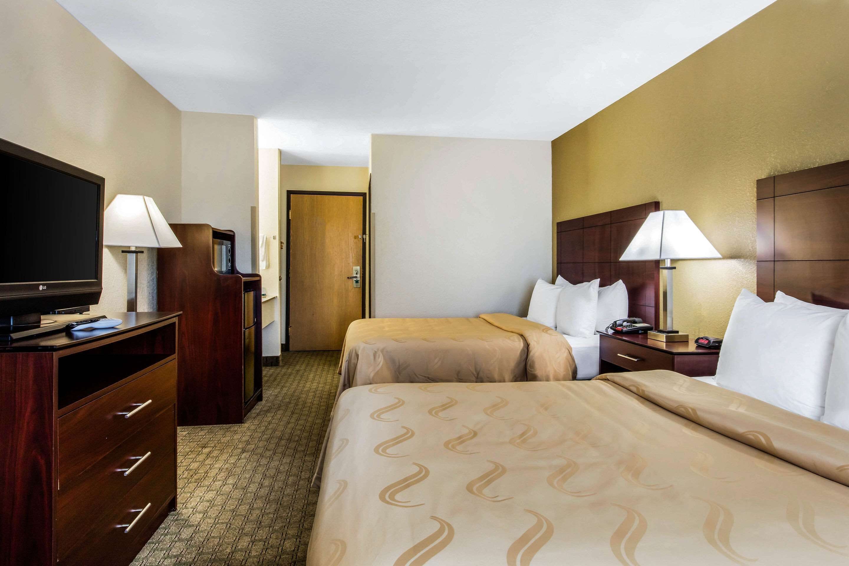 Quarto de hotel com duas camas, televisão, escrivaninha com luminária e guarda-roupa.