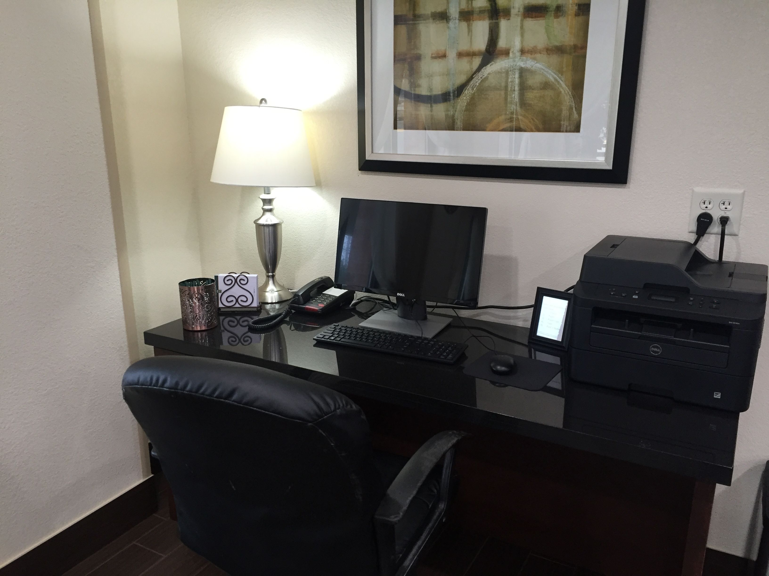 A hotel desk with a computer, printer, lamp, and a framed picture on the wall.