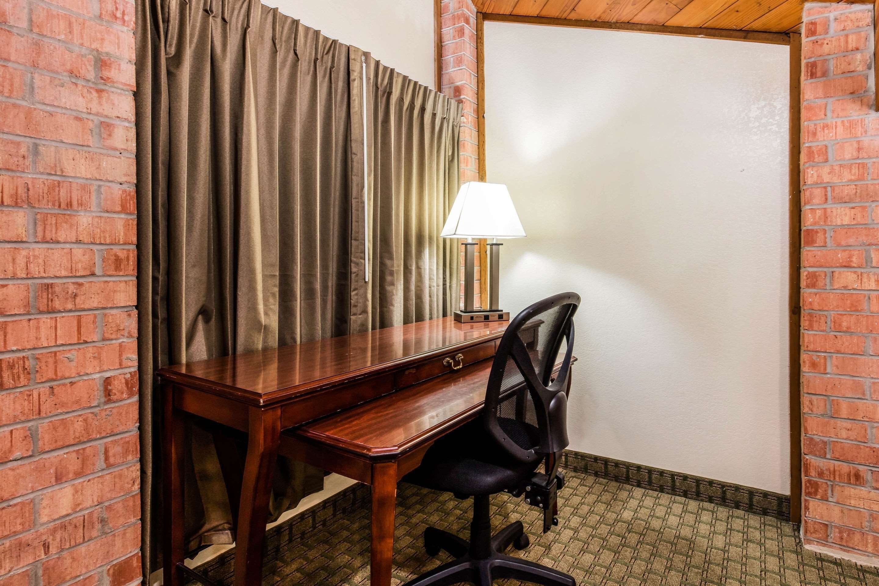 Habitación de esquina con un escritorio de madera, una silla de oficina negra, una lámpara y una pared de ladrillo.