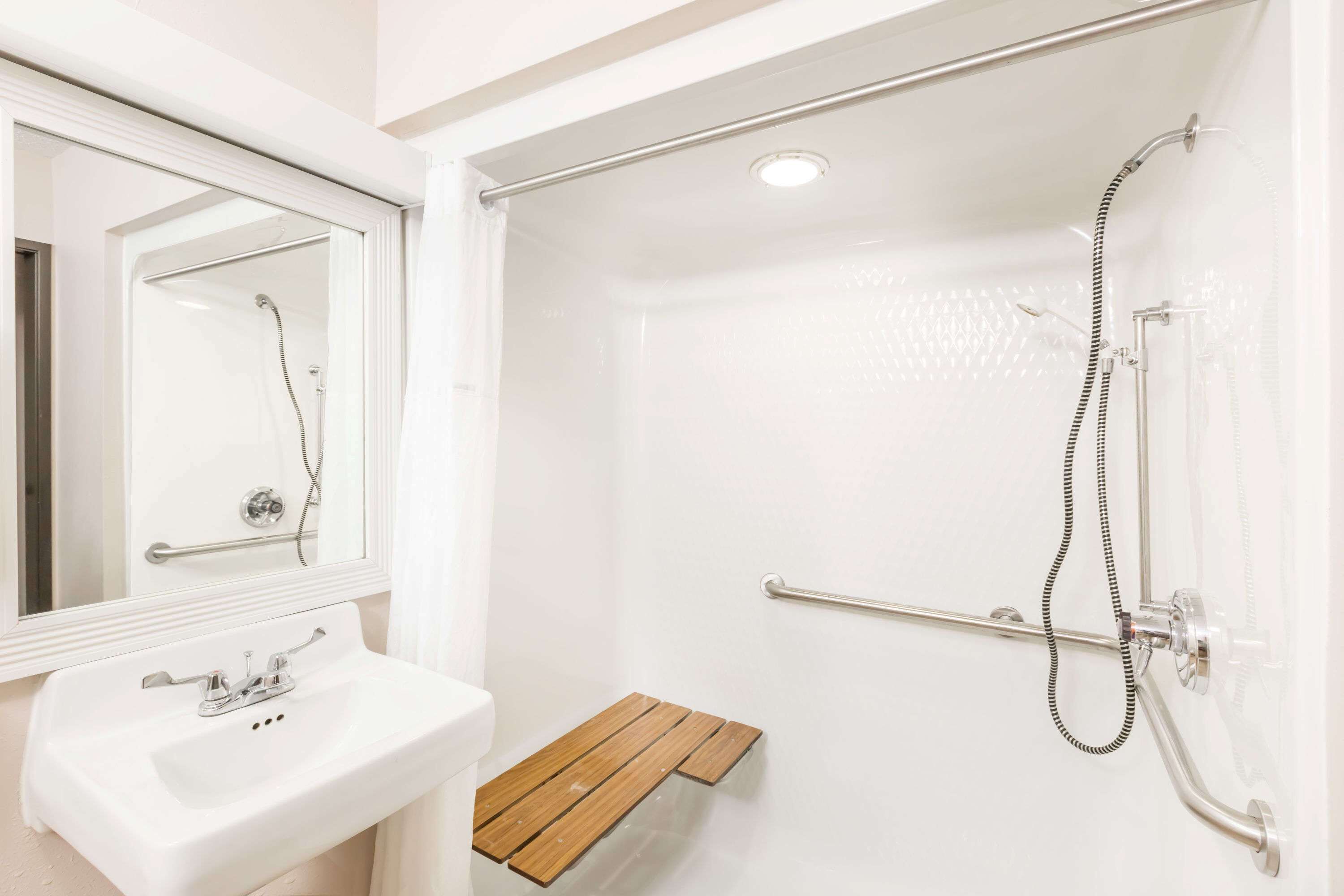 Un baño con un lavabo blanco, un espejo, una ducha con cabezal de ducha manual y un estante de madera.