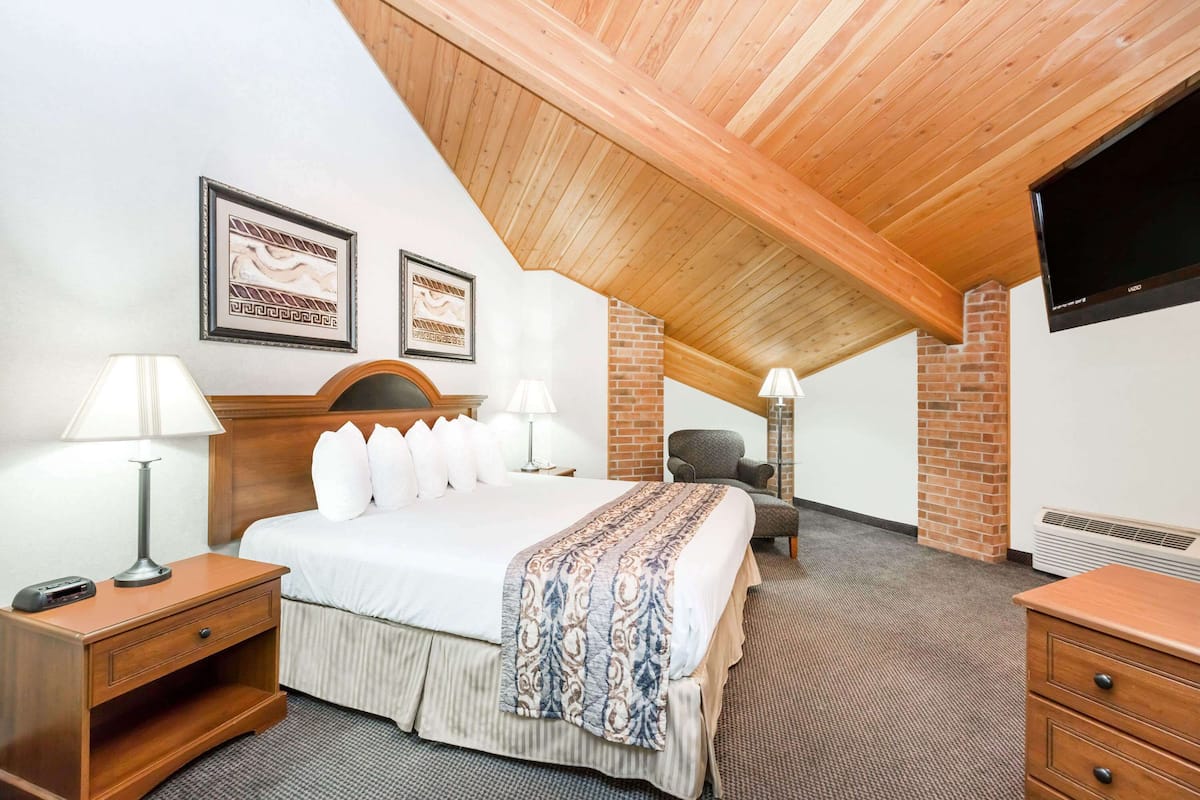 Une chambre d’hôtel avec un plafond en pente, un lit avec une literie blanche et un couvre-lit à motifs, deux lampes de chevet, un téléviseur à écran plat fixé au mur et une cheminée en briques.