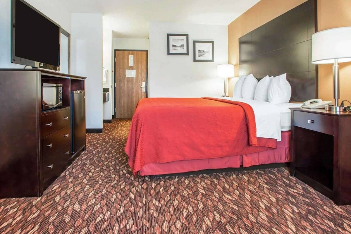 A hotel room with a bed, a desk with a lamp, a television on a dresser, and a door leading to another room.