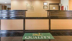 Un área de recepción con mostrador, un cartel en la pared que dice "QUALITY INN & SUITES BY CHOICE HOTELS" y un felpudo verde con el logotipo del hotel.
