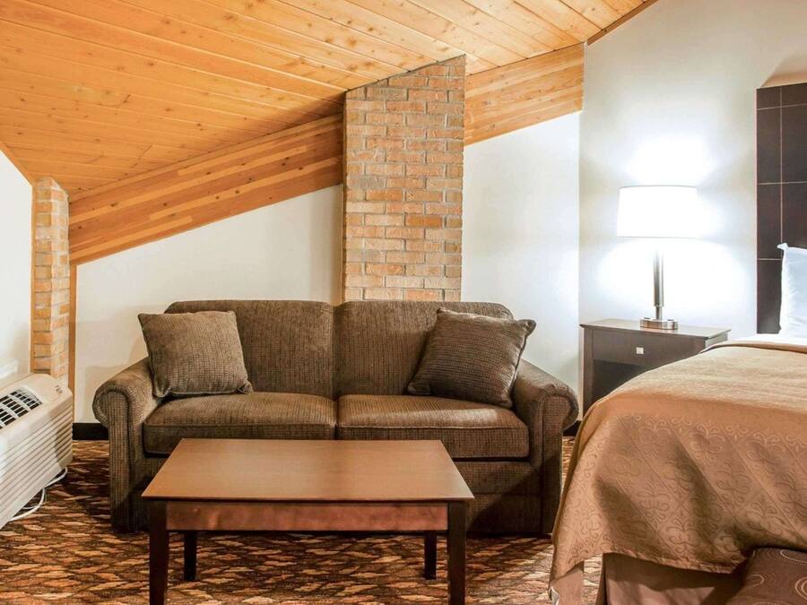 A cozy room with a wooden ceiling, a brick wall, a sofa, a small table, and a bed with a patterned bedspread.