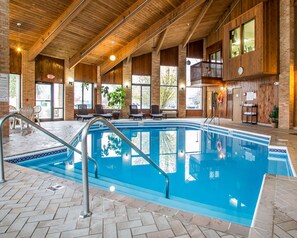 Piscina interior con techo de madera y paredes de ladrillo, rodeada de sillones de descanso y vistas al exterior.