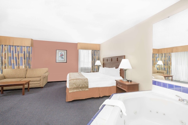 A hotel room with a bed, a sofa, a small table, a nightstand, a lamp, a mirror, and a bathtub.