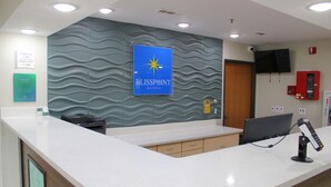 A hotel reception area with a check-in/out sign, a Blisspoint Hotels sign, a flat-screen TV, a fire extinguisher, and a self-service kiosk. - Blisspoint Inn Columbia City (Columbia City)