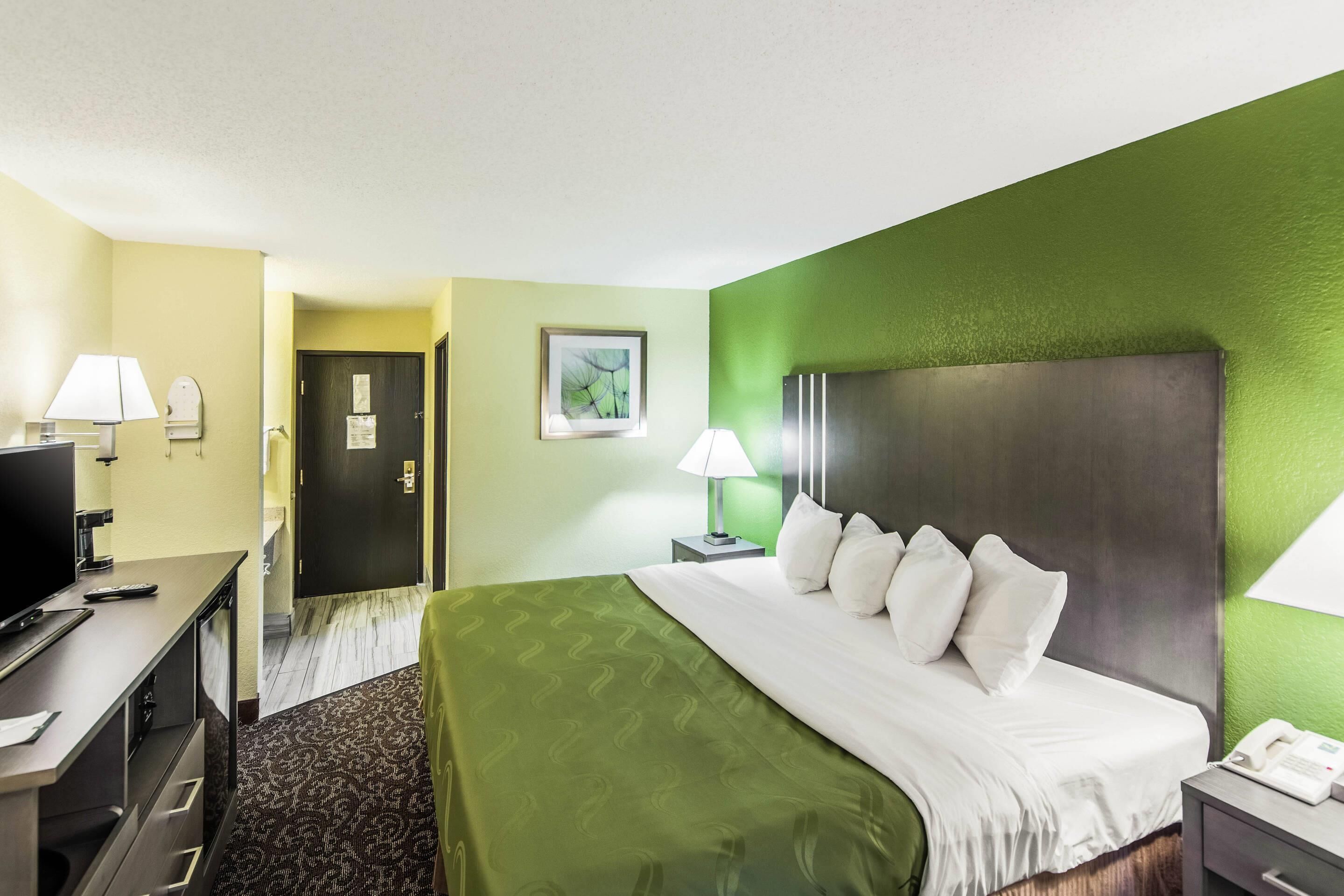 Habitación de hotel con cama, mesita de noche, lámpara y televisor sobre un escritorio.