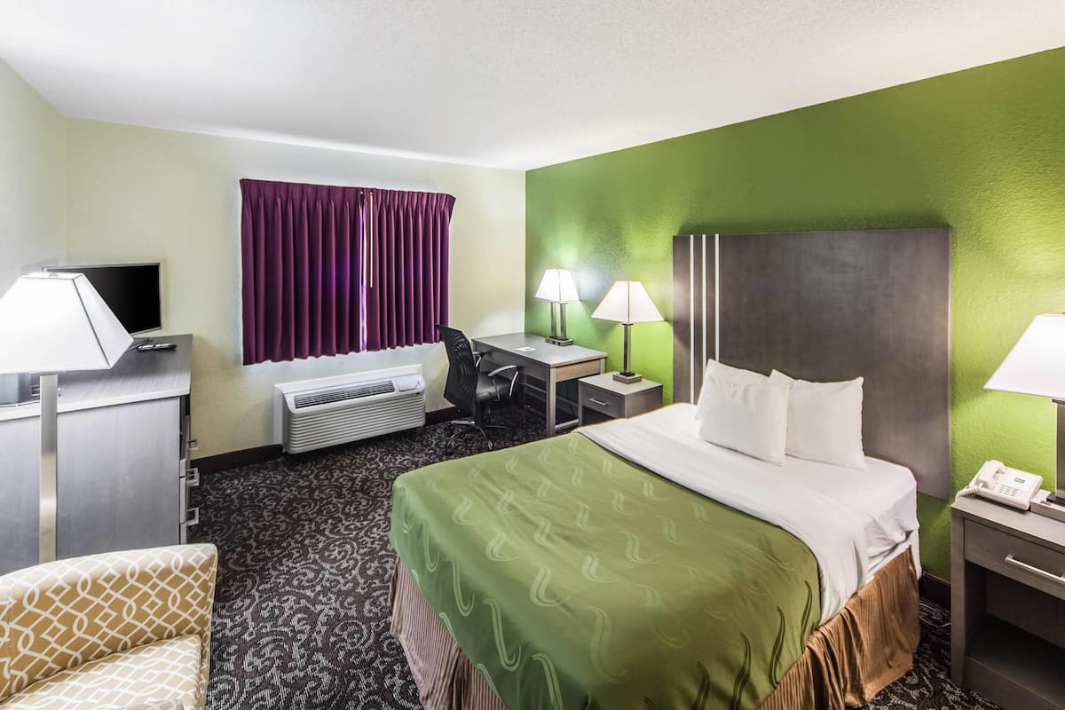 A hotel room with a bed, desk, chair, air conditioning unit, and a television.