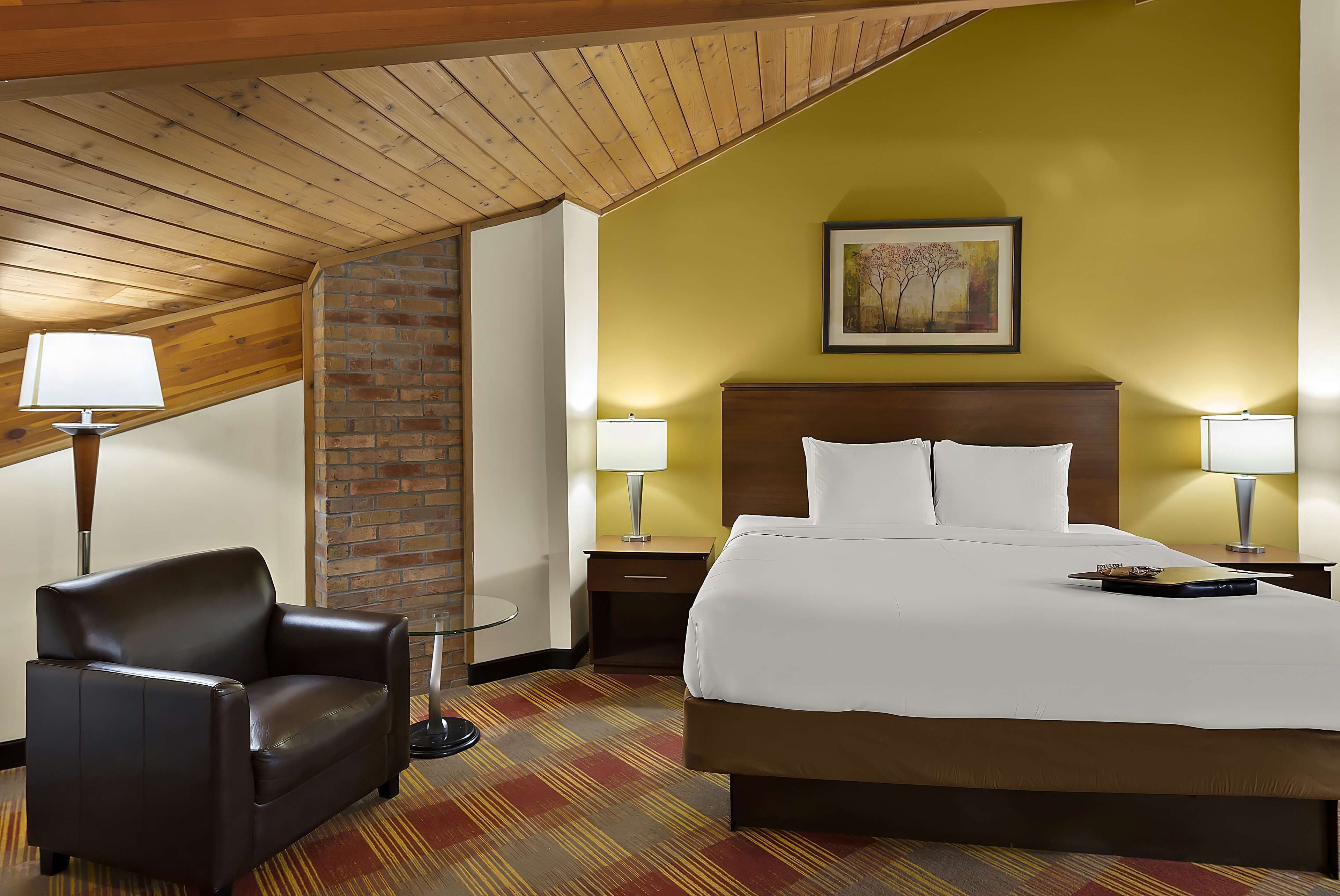 A hotel room with a large bed, two bedside tables with lamps, a leather armchair, and a brick wall.