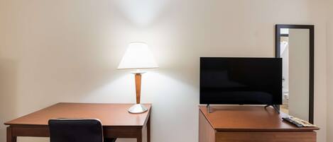 Habitación de hotel con escritorio de madera, una silla negra, lámpara, televisor, espejo y cómoda con cajones.