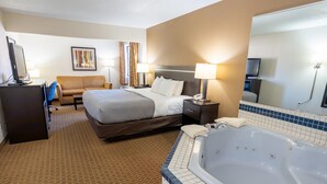 Una habitación de hotel con una cama grande, un escritorio con computadora, un sofá, un televisor, un baño con bañera y un espejo grande.