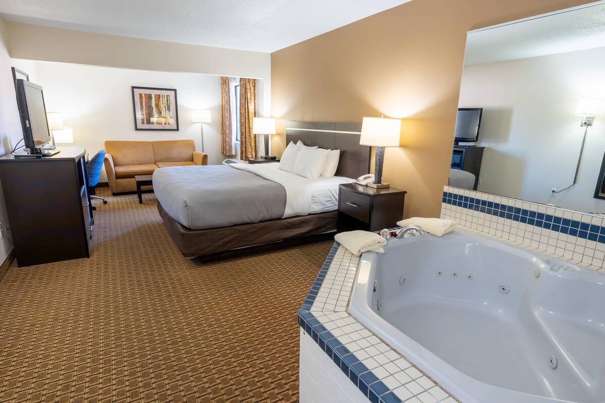 A hotel room with a large bed, a desk with a computer, a sofa, a TV, a bathroom with a bathtub, and a large mirror.