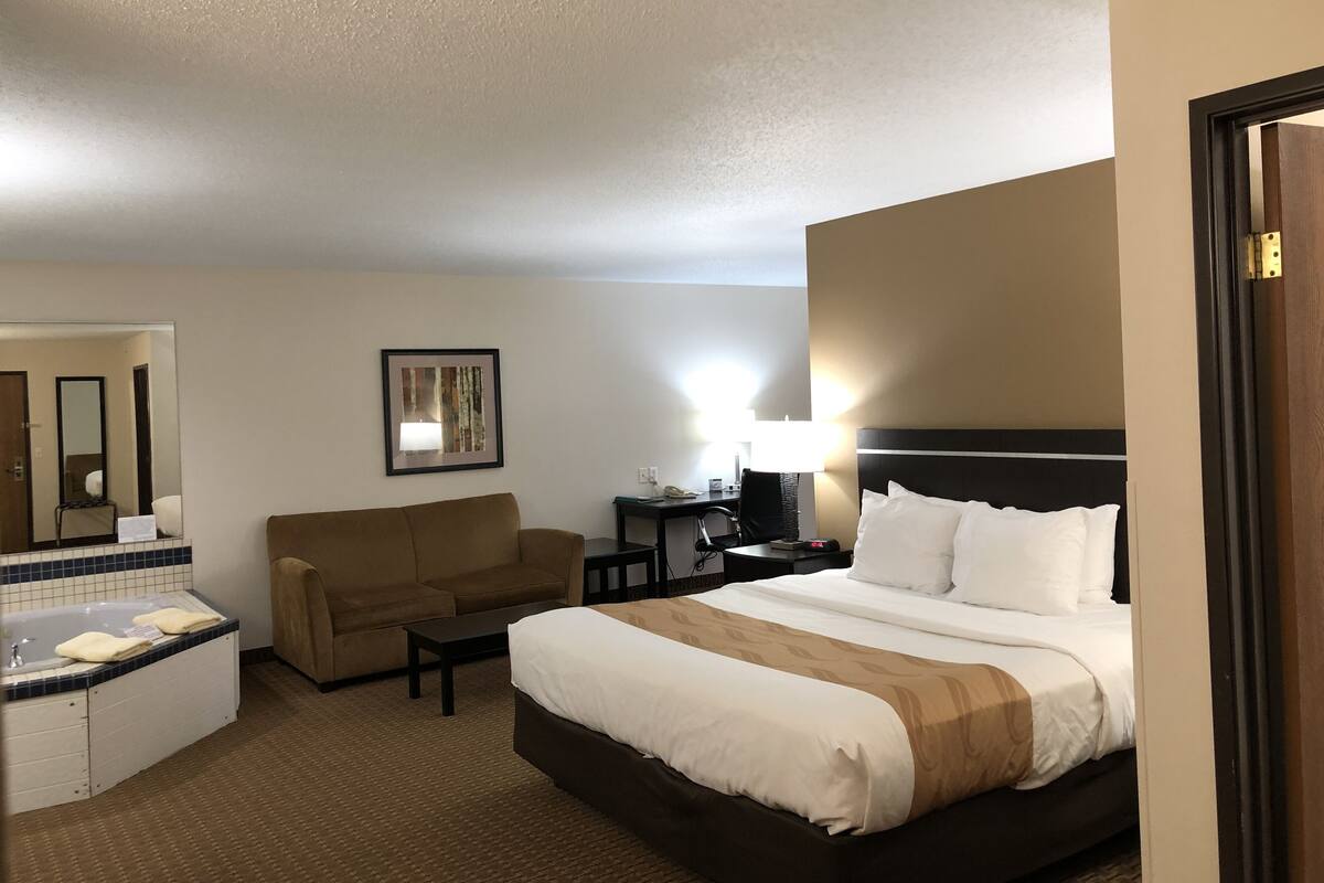 A hotel room with a bed, a sofa, a desk, a chair, a lamp, and a bathroom with a bathtub and a mirror.