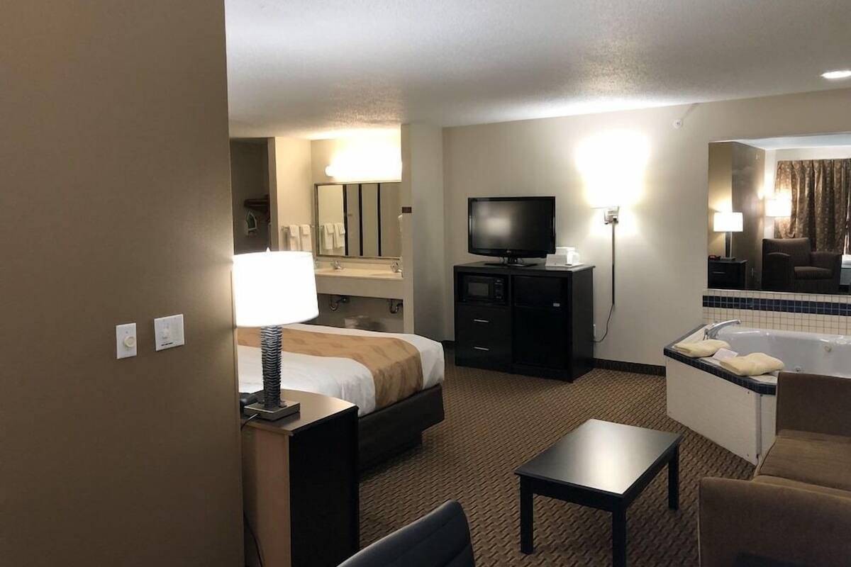 A hotel room with a bed, a desk with a lamp, a TV, a desk lamp, a chair, a small table, a bathroom with a bathtub, and a mirror.