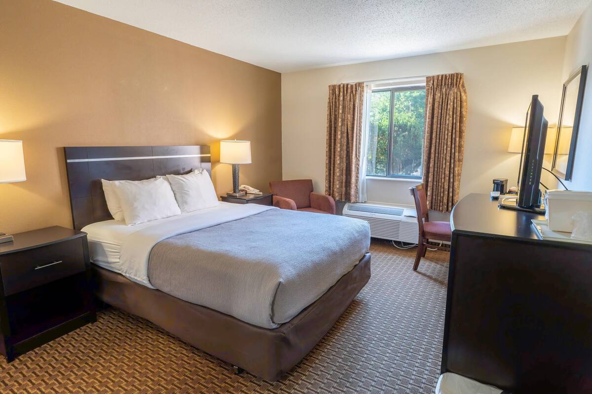 A hotel room with a large bed, two bedside tables with lamps, a desk with a computer, a chair, and a window with curtains.