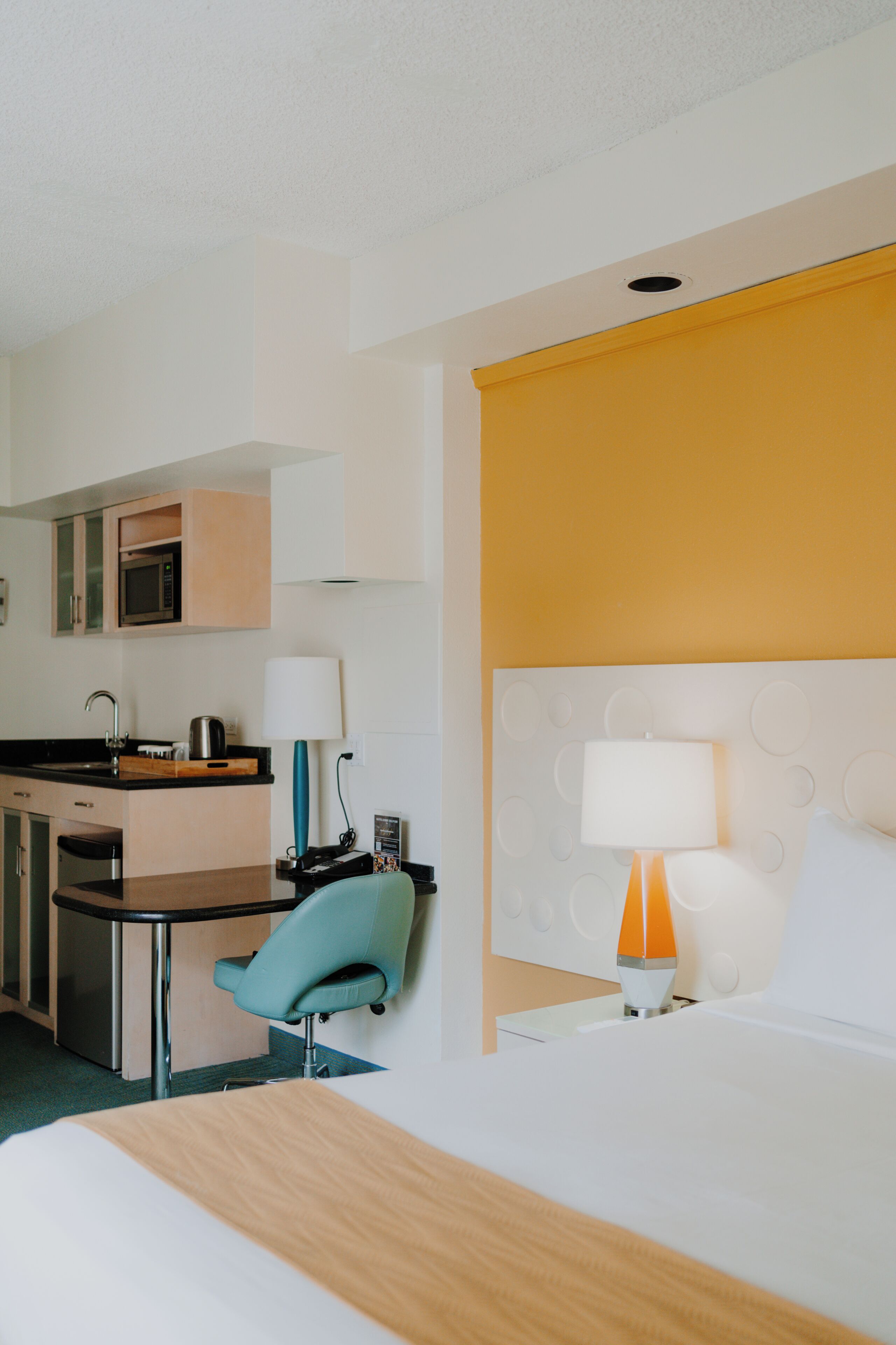 A hotel room with a bed, a desk, a chair, a microwave, and a lamp.
