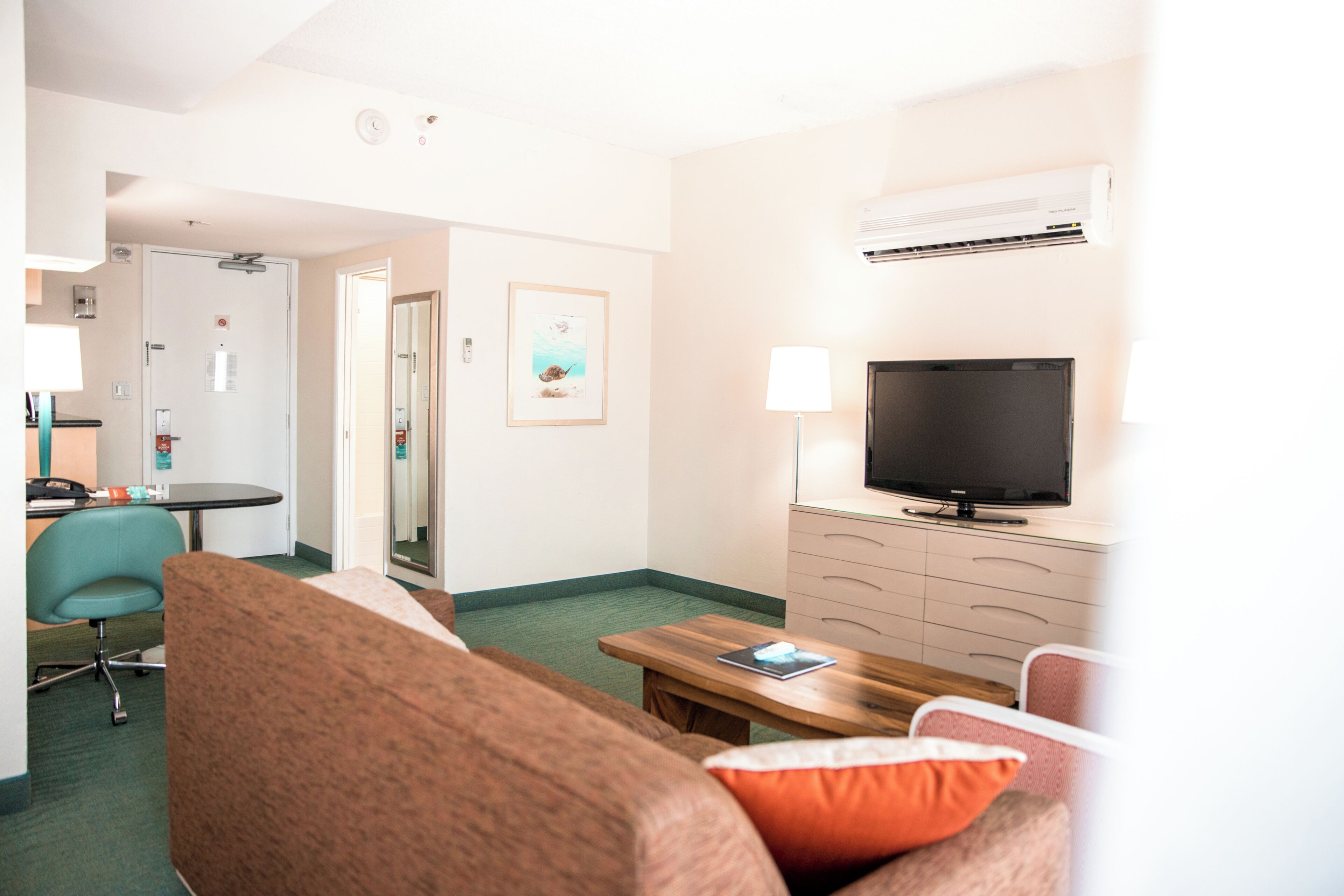 A hotel room with a TV, a wooden table, a couch, a chair, a desk, and a door.