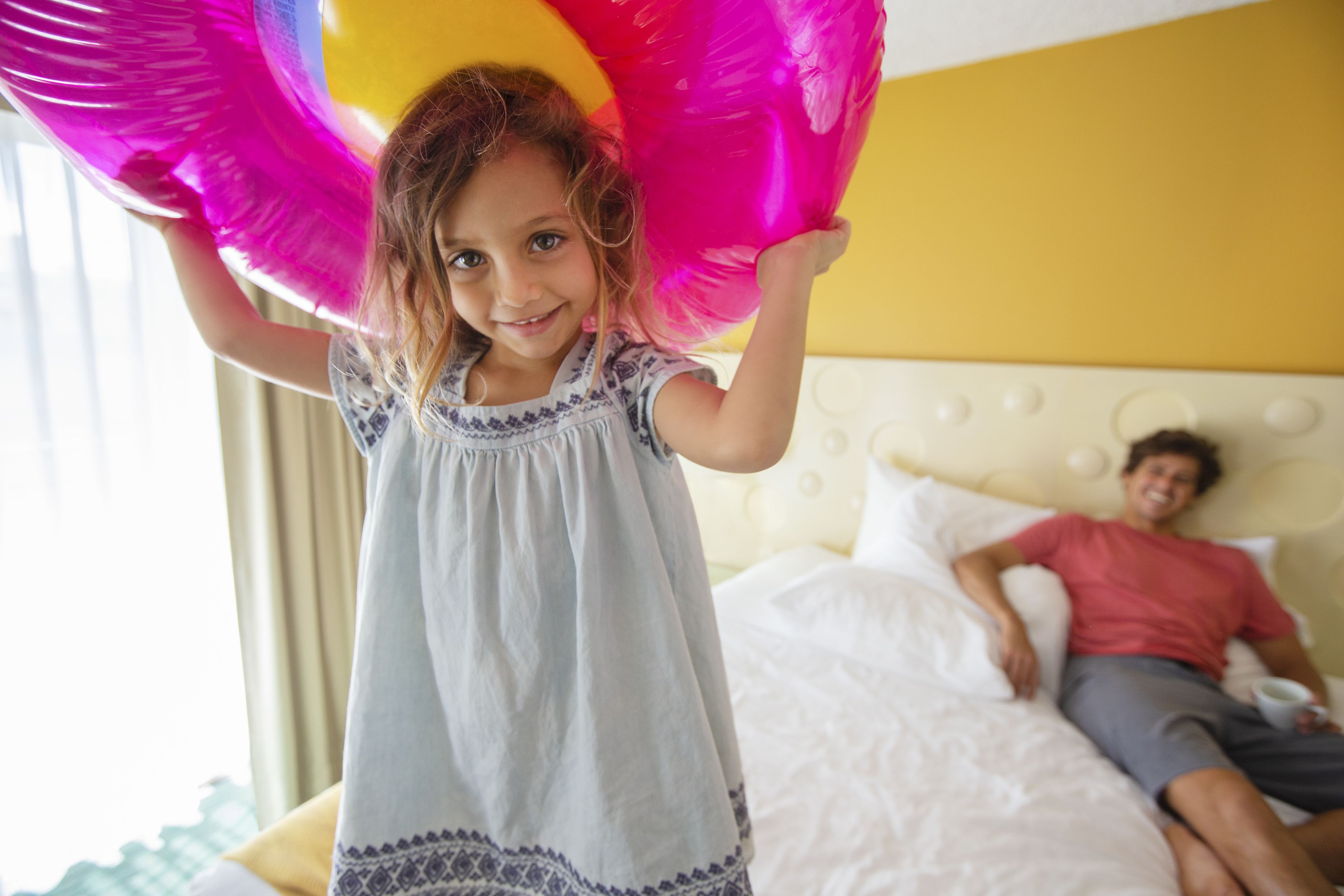 A child holding a large pink balloon in a room with a bed and a person lying on it.