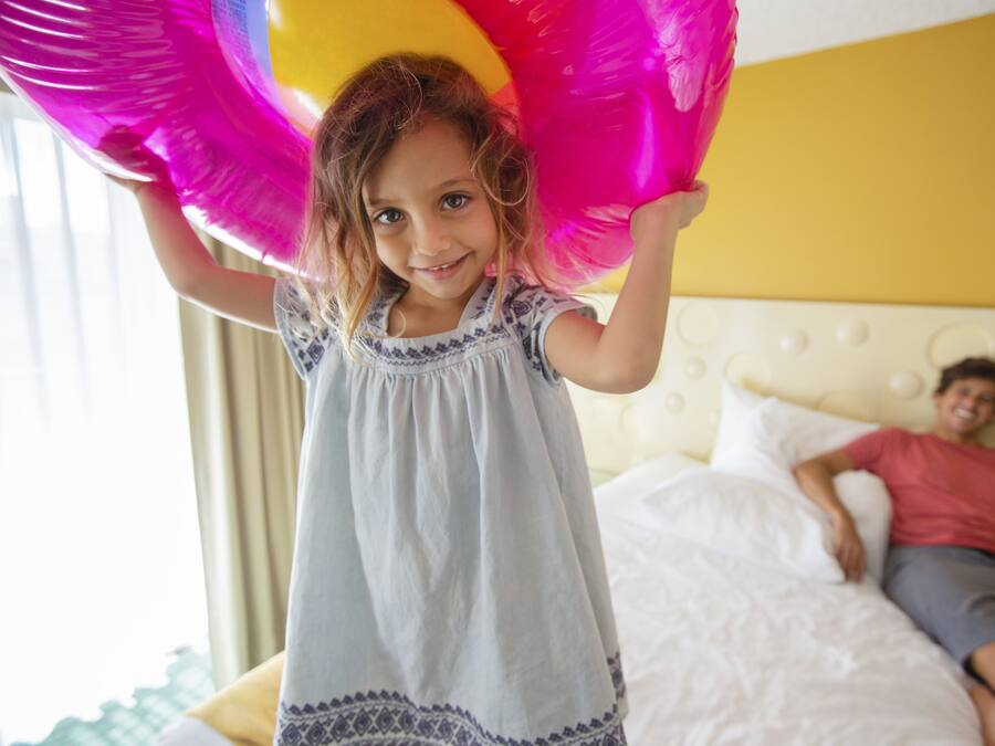 A child holding a large pink balloon in a room with a bed and a person lying on it.