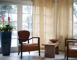 Una habitación con un ventanal grande, una planta en maceta, una silla de madera, una mesa de madera y un cuenco.