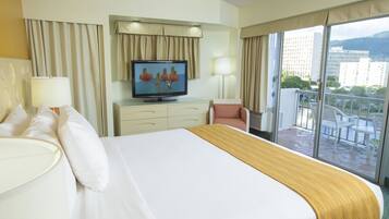 A hotel room with a large bed, a television, a desk, and a balcony with a view of buildings.