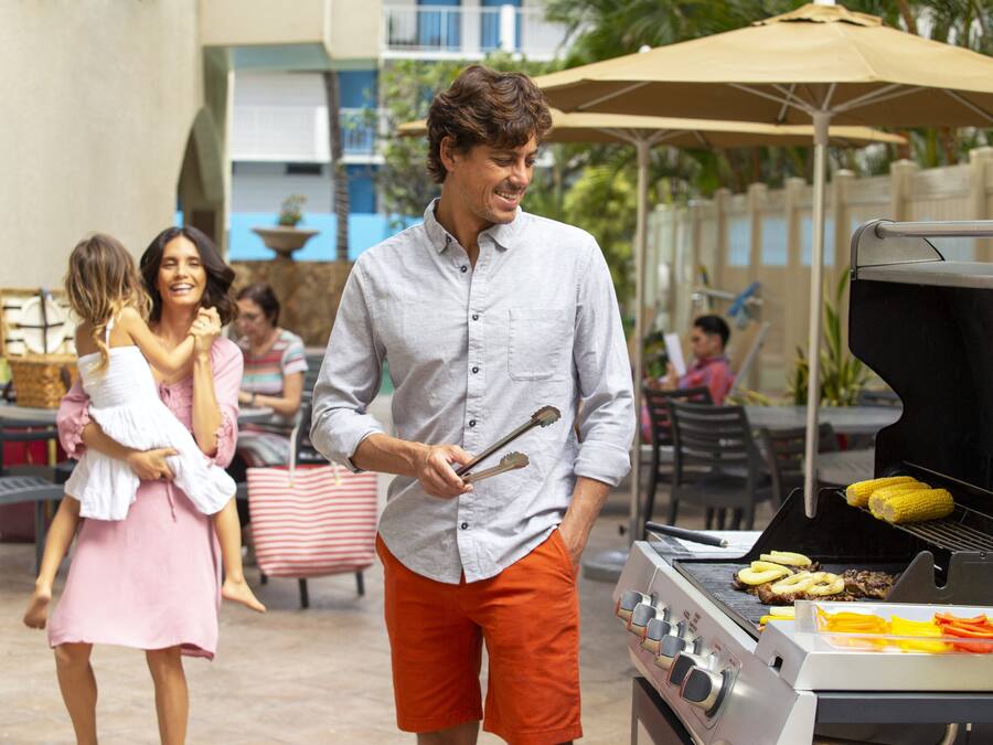 A man grilling food outdoors with a grill and tongs, a dining area with tables and chairs, and a building in the background.