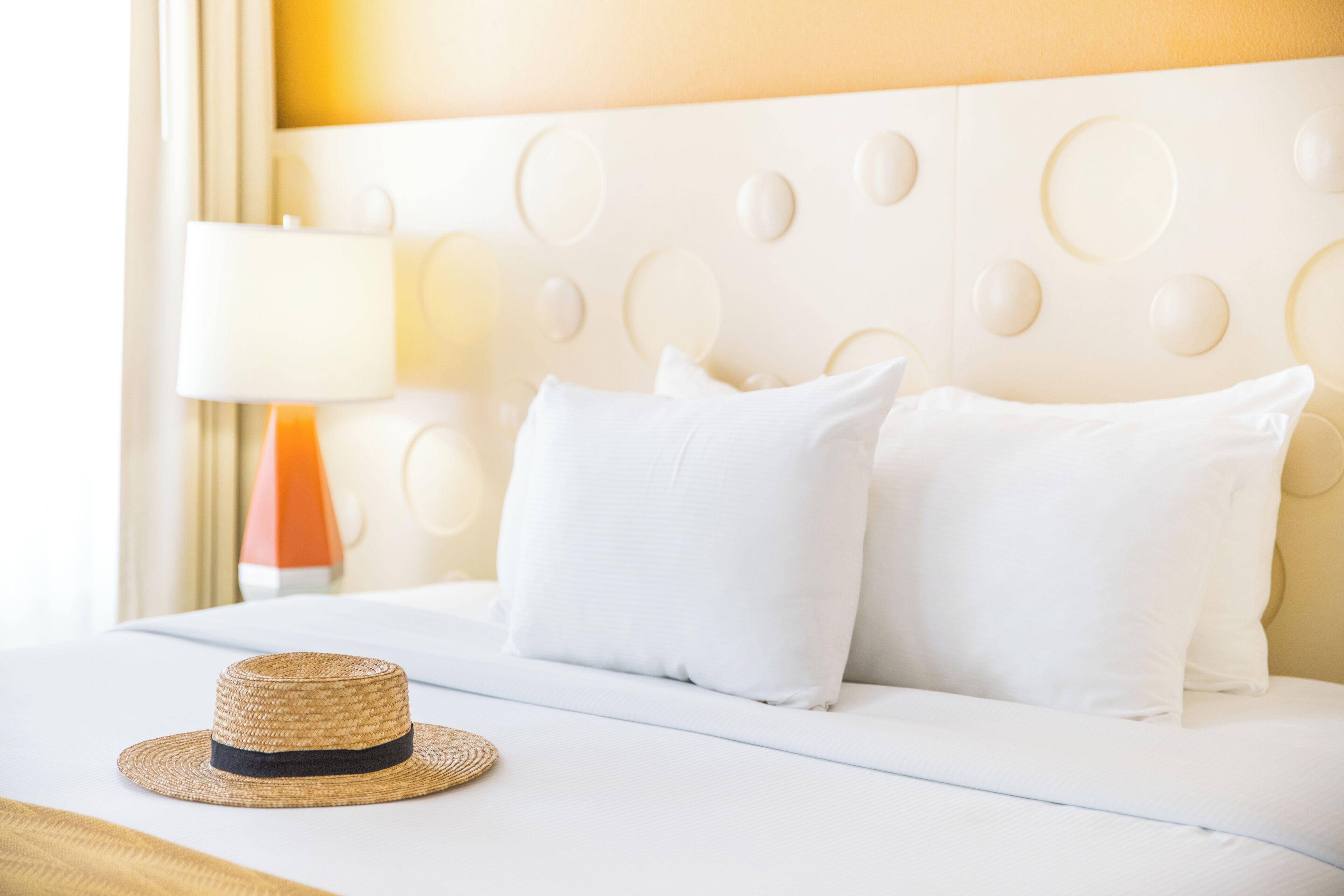 A hotel room with a white bed, a straw hat, and a lamp.
