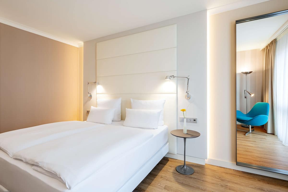 Ein modernes Hotelzimmer mit einem großen Bett, Nachttischlampen, einem kleinen Tisch mit einer Vase sowie einem Spiegel, der einen blauen Sessel und Vorhänge reflektiert.