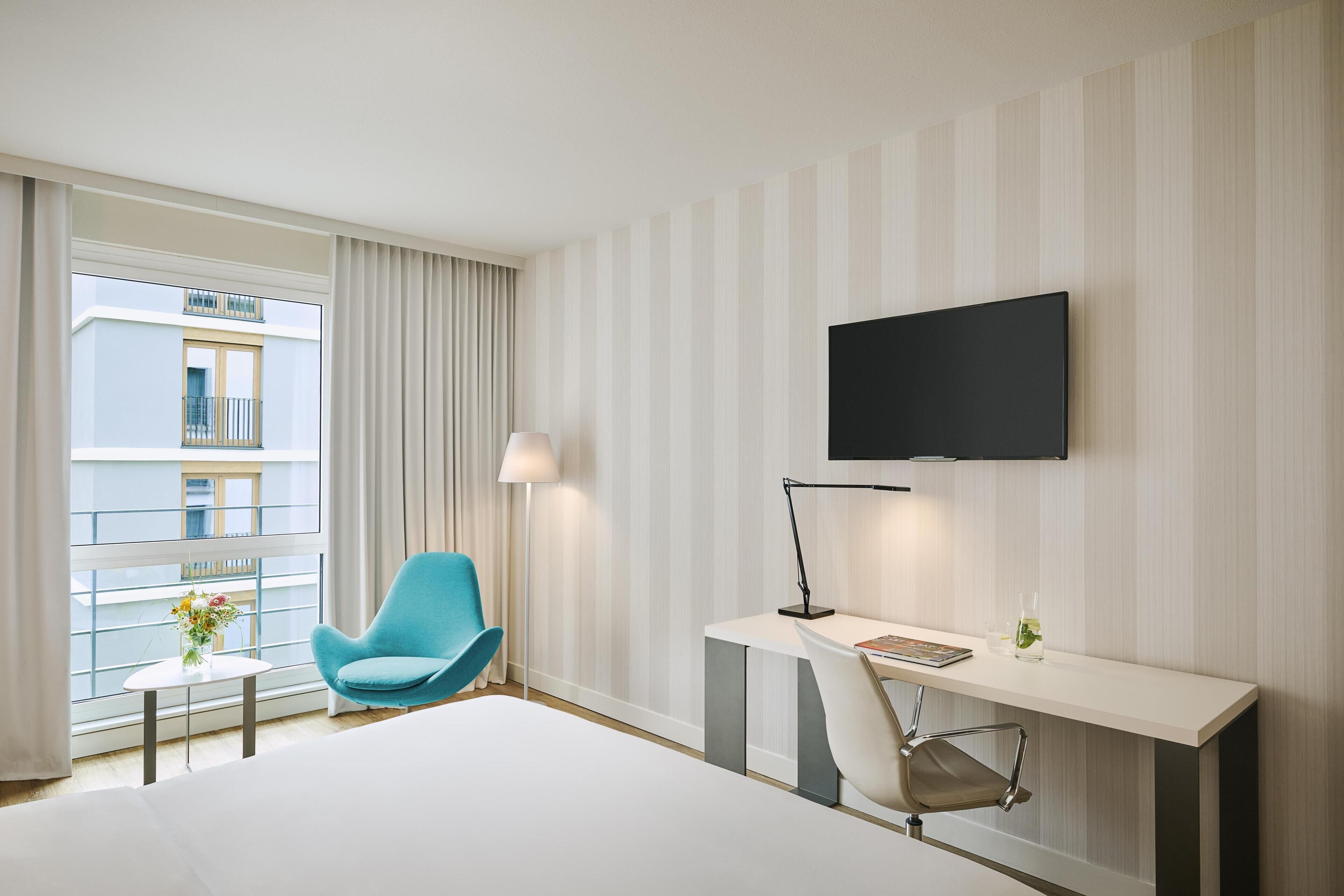 Habitación de hotel con cama, escritorio, silla, televisor y ventana con cortinas.