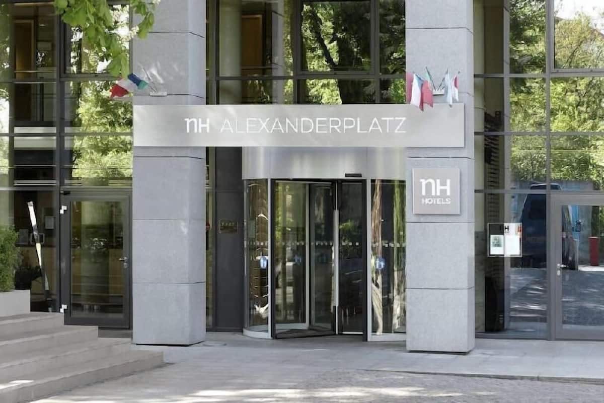 Ingången till ett hotell med en fasad av glas, en skylt med texten ”NH ALEXANDERPLATZ” och flaggor på vänster sida.