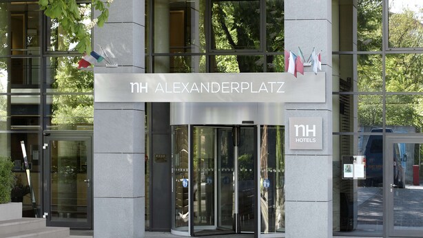 La entrada del hotel NH Alexanderplatz, con fachada de vidrio moderna, una puerta giratoria y un letrero que indica el nombre del hotel.