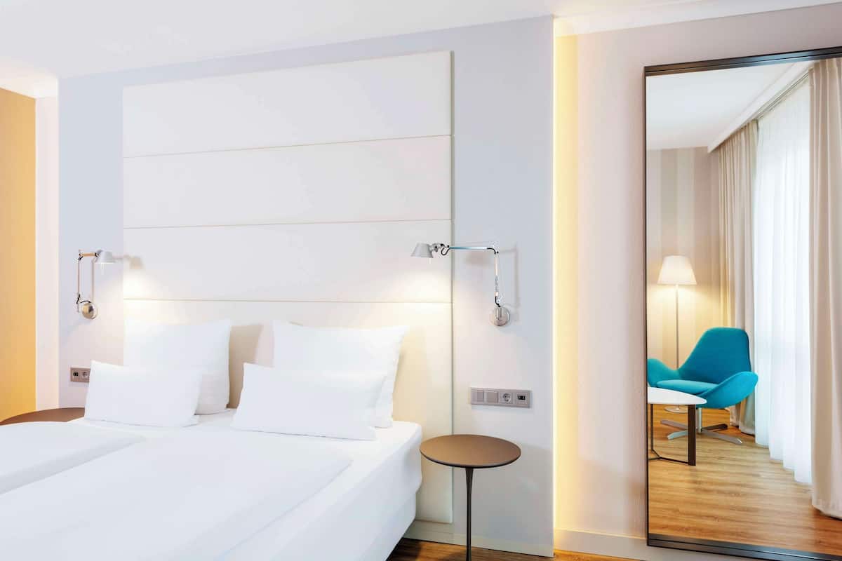 Ein Hotelzimmer mit einem großen Bett, Nachttischen, einem Spiegel und einem Blick auf einen blauen Stuhl und Vorhänge im Spiegelbild.