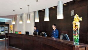 A reception desk with two staff members, a clock, and a vase of flowers. - NH Frankfurt Airport West (Raunheim)