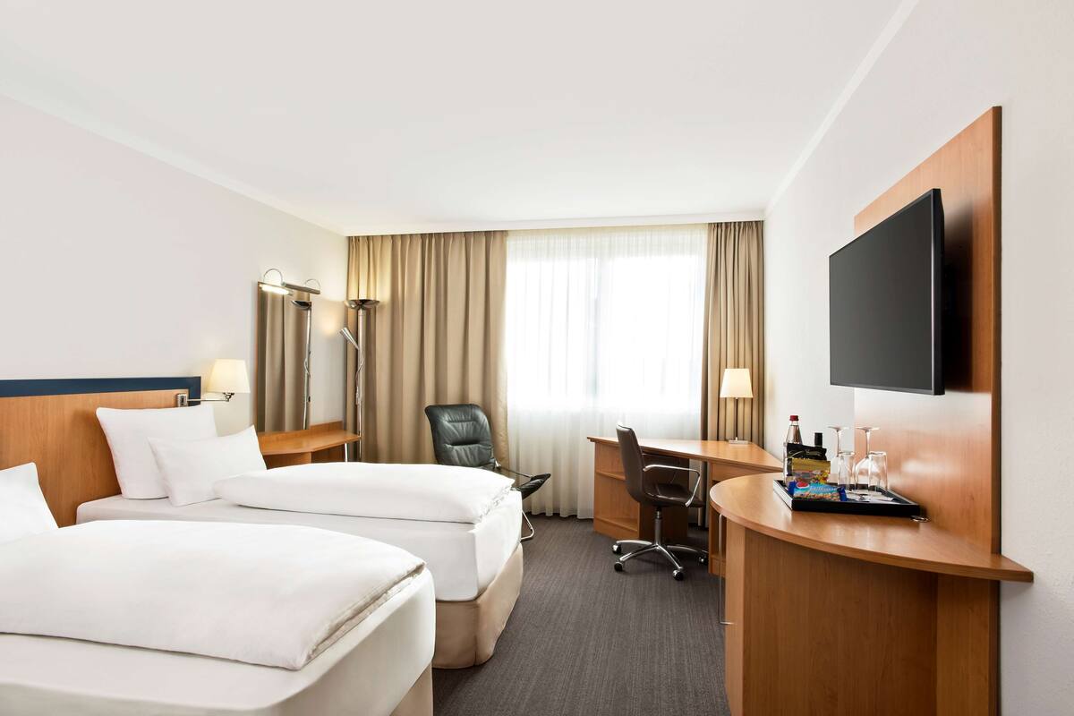 Quarto de hotel com duas camas, uma escrivaninha, uma cadeira, televisão e janela com cortinas.