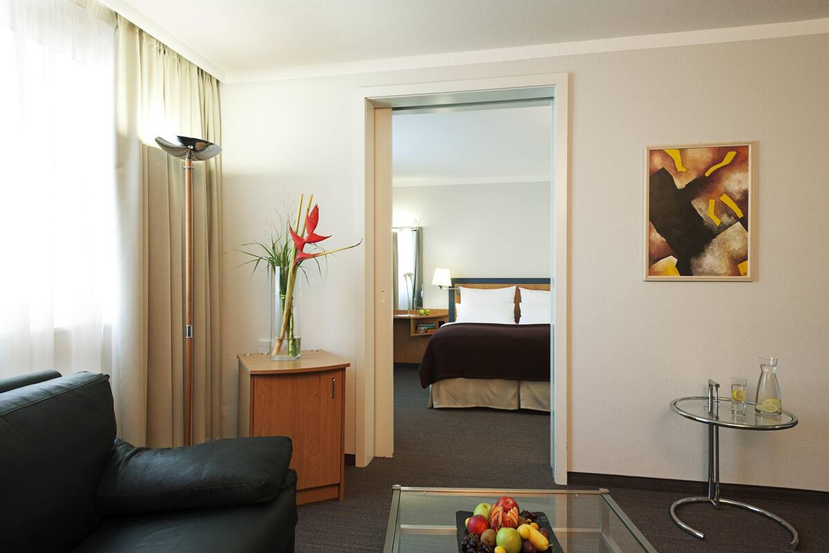 Une chambre d’hôtel avec un lit, un coin salon avec un canapé, une petite table avec des fruits, une lampe et un tableau au mur.