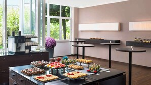 A modern kitchen with a buffet setup, large windows, and a coffee station. - NH Frankfurt Airport West (Raunheim)