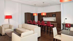 A modern living room with a sofa set, a dining table with chairs, a red lamp, and a kitchen area with bar stools. - NH Frankfurt Airport West (Raunheim)