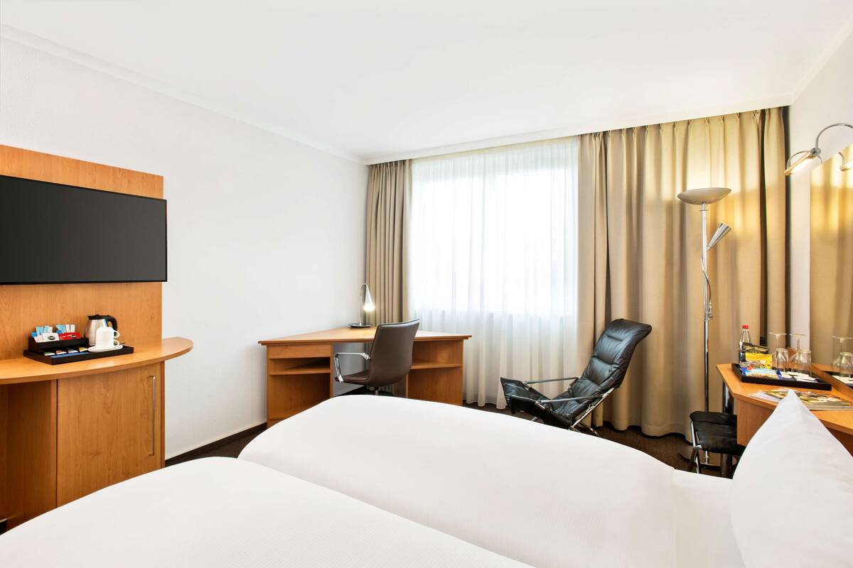 Quarto de hotel com cama, escrivaninha, cadeira, televisão fixada na parede e janela com cortinas.