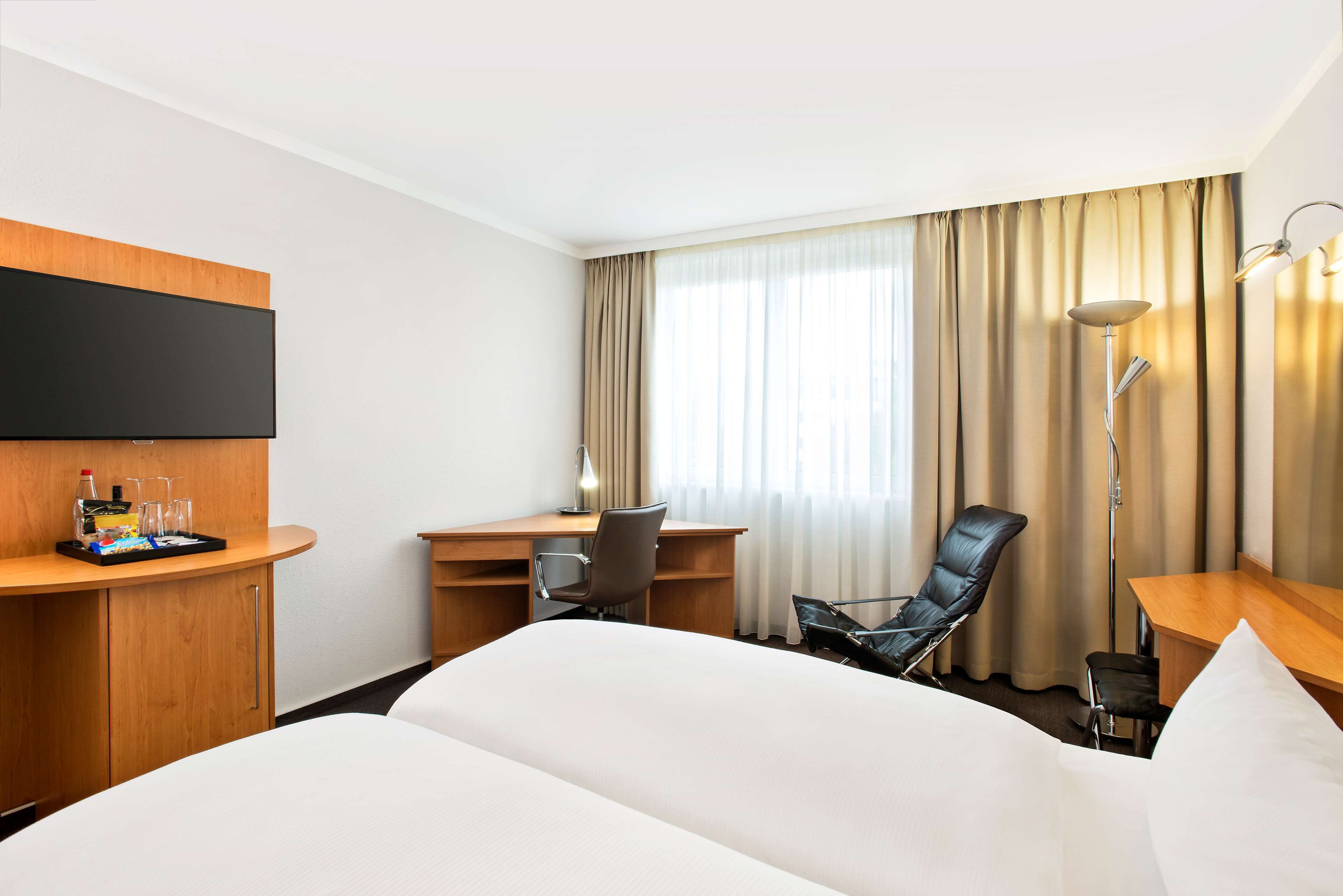 A hotel room with a large bed, a desk with a chair, a TV mounted on the wall, and a window with curtains.