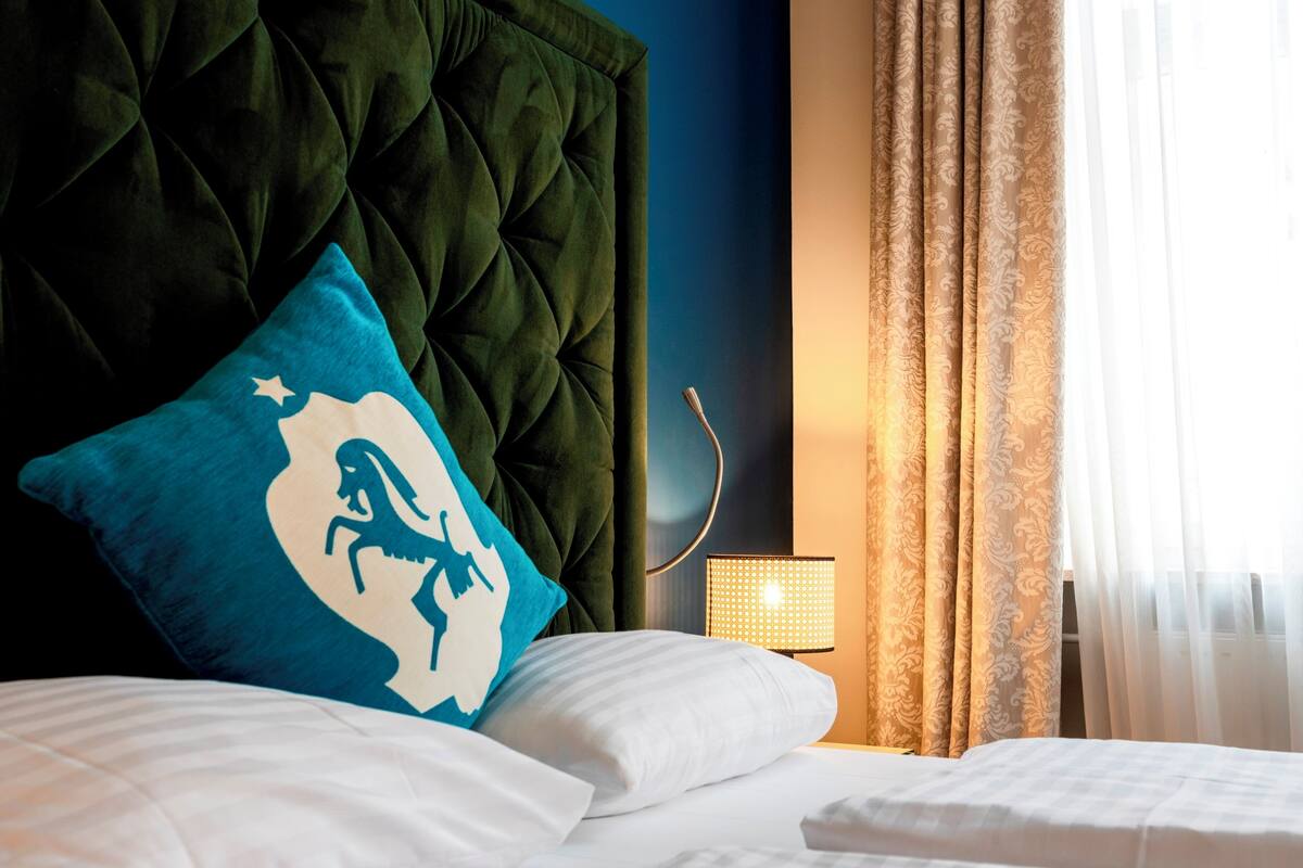 A neatly made bed with white linens, a blue pillow with a white design, a green upholstered headboard, a bedside lamp, and a window with sheer curtains.