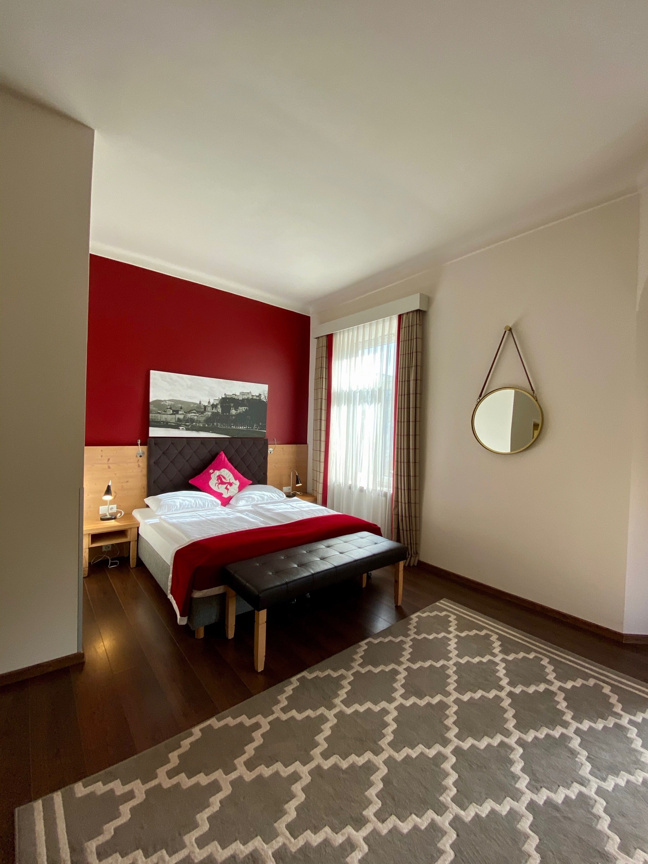 A hotel room with a bed, a bedside table with a lamp, a bench, a large mirror, and a patterned rug.