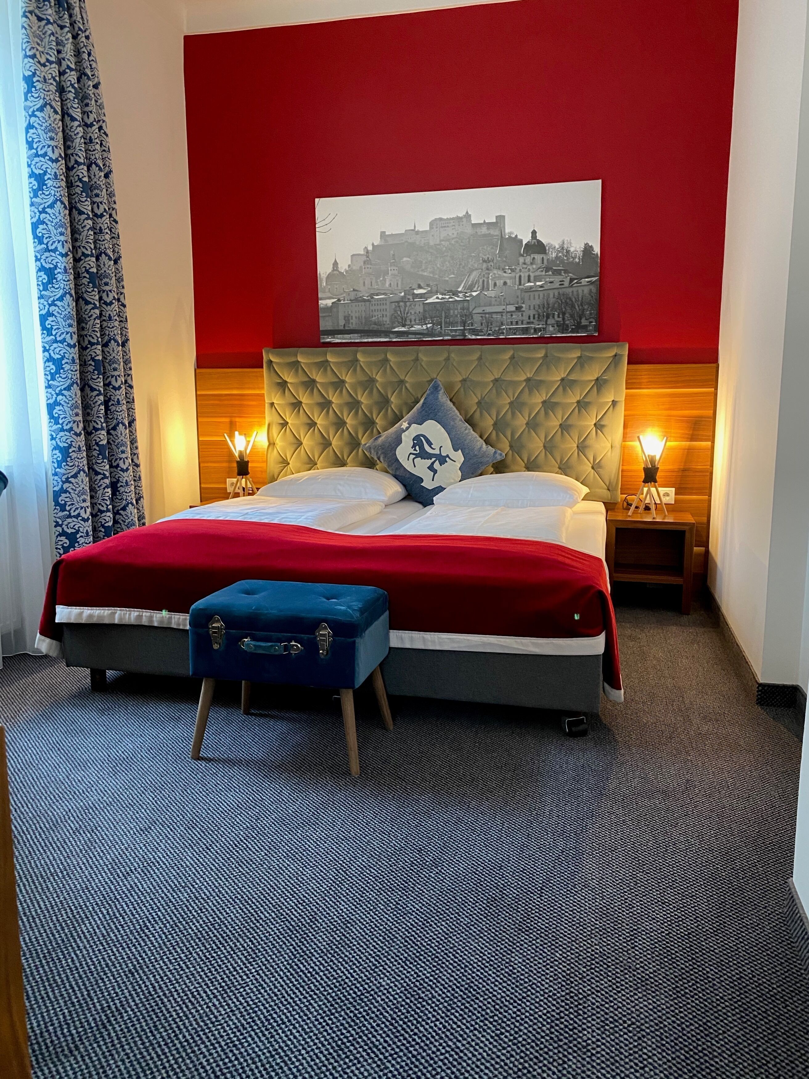 Ein Hotelzimmer mit einem großen Bett, roten Wänden, einem Stadtbildgemälde, zwei Nachttischlampen und einem blauen Koffer.